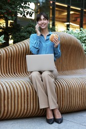 Photo of Lunch time. Happy businesswoman with hamburger and laptop talking on smartphone on bench outdoors