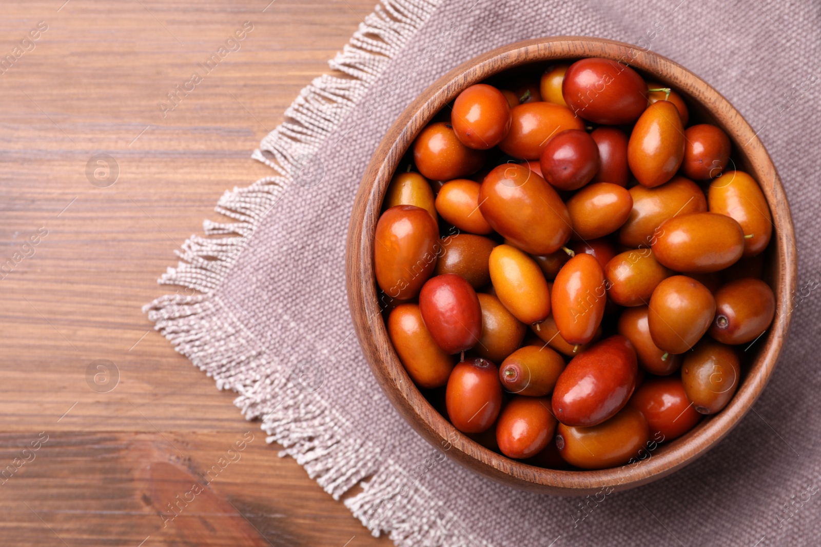 Photo of Fresh Ziziphus jujuba fruits in bowl with napkin on wooden table, top view. Space for text