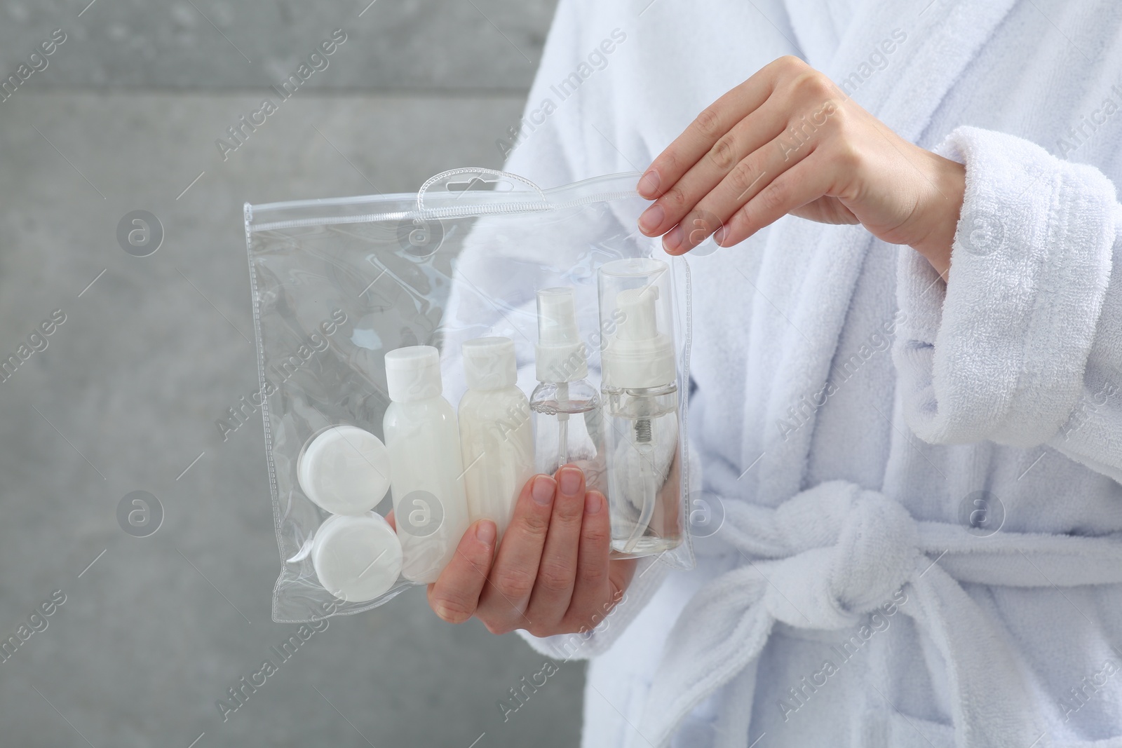 Photo of Woman holding plastic bag of cosmetic travel kit against grey wall, closeup. Bath accessories