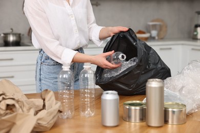 Photo of Garbage sorting. Woman putting plastic bottle into bag at wooden table indoors, closeup
