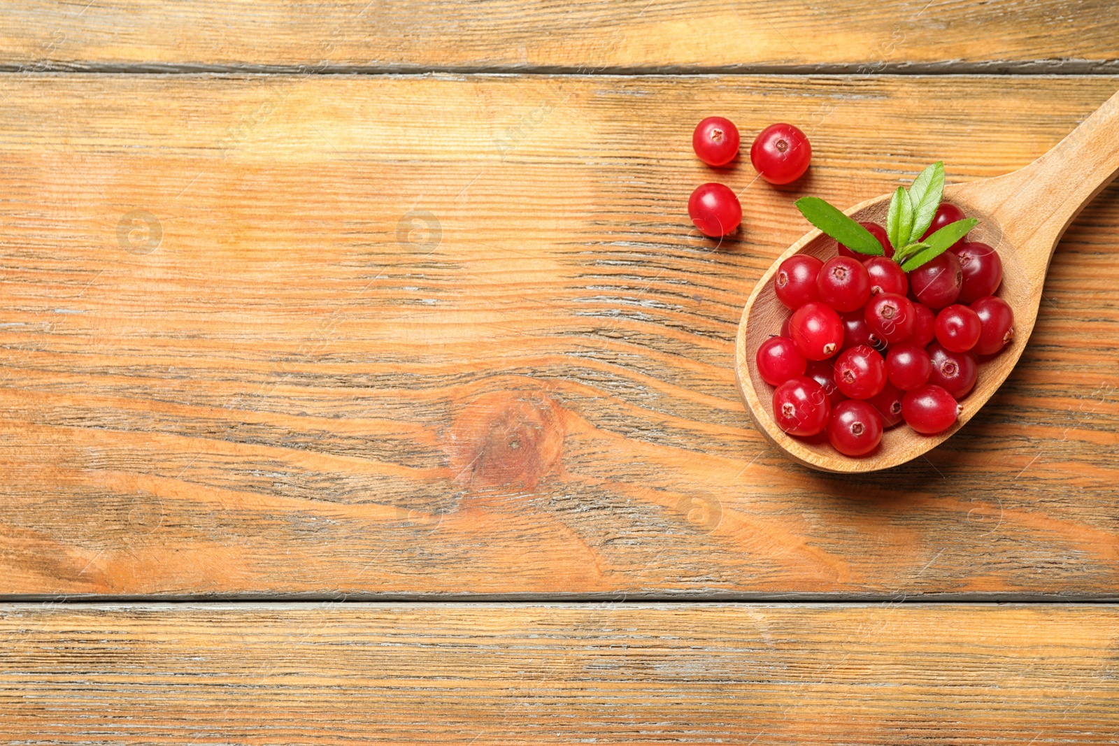 Photo of Spoon with tasty cranberries and green leaves on wooden table, flat lay. Space for text