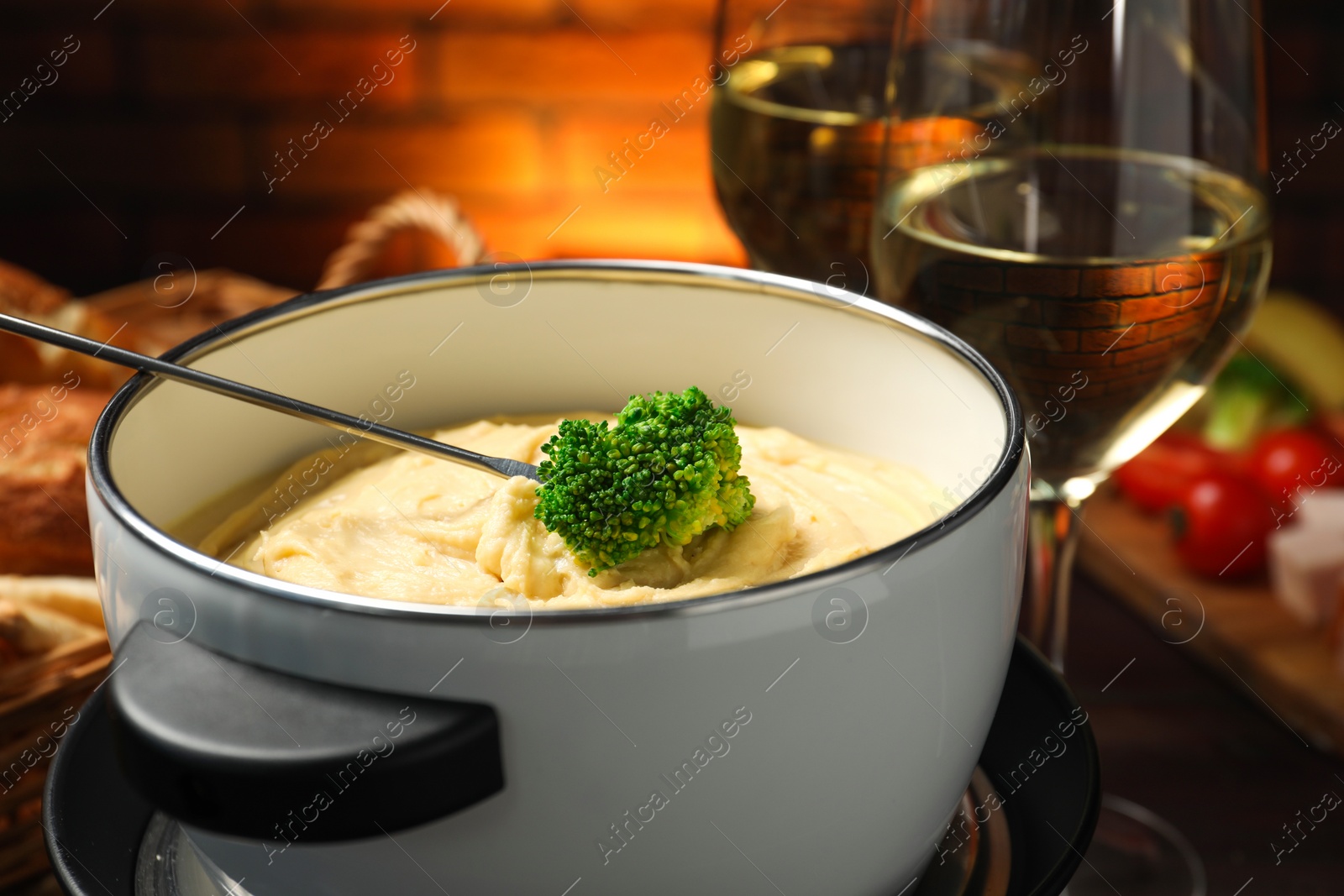 Photo of Dipping piece of broccoli into fondue pot with melted cheese at table, closeup