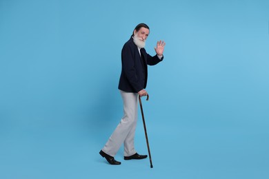 Senior man with walking cane waving on light blue background