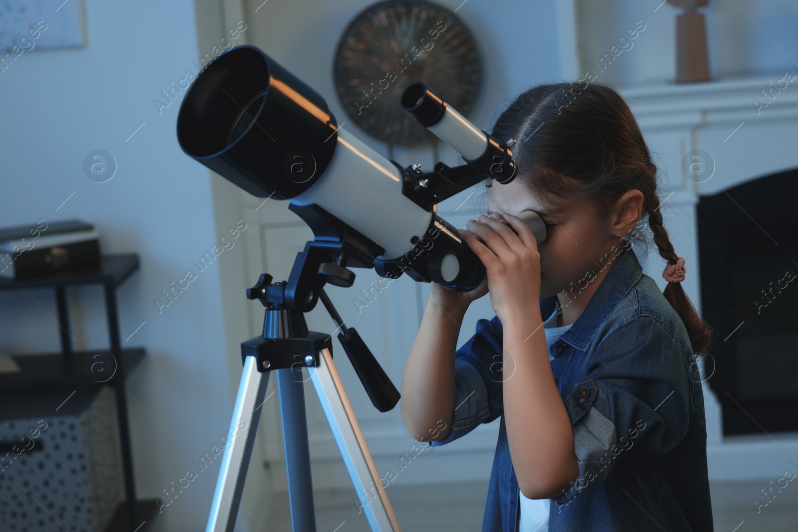 Photo of Cute little girl looking at stars through telescope in room