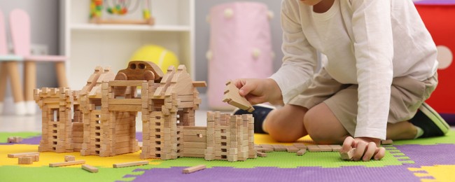 Photo of Little boy playing with wooden construction set on puzzle mat in room, closeup. Child's toy