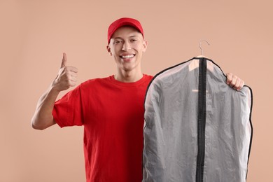 Photo of Dry-cleaning delivery. Happy courier holding garment cover with clothes and showing thumbs up on beige background