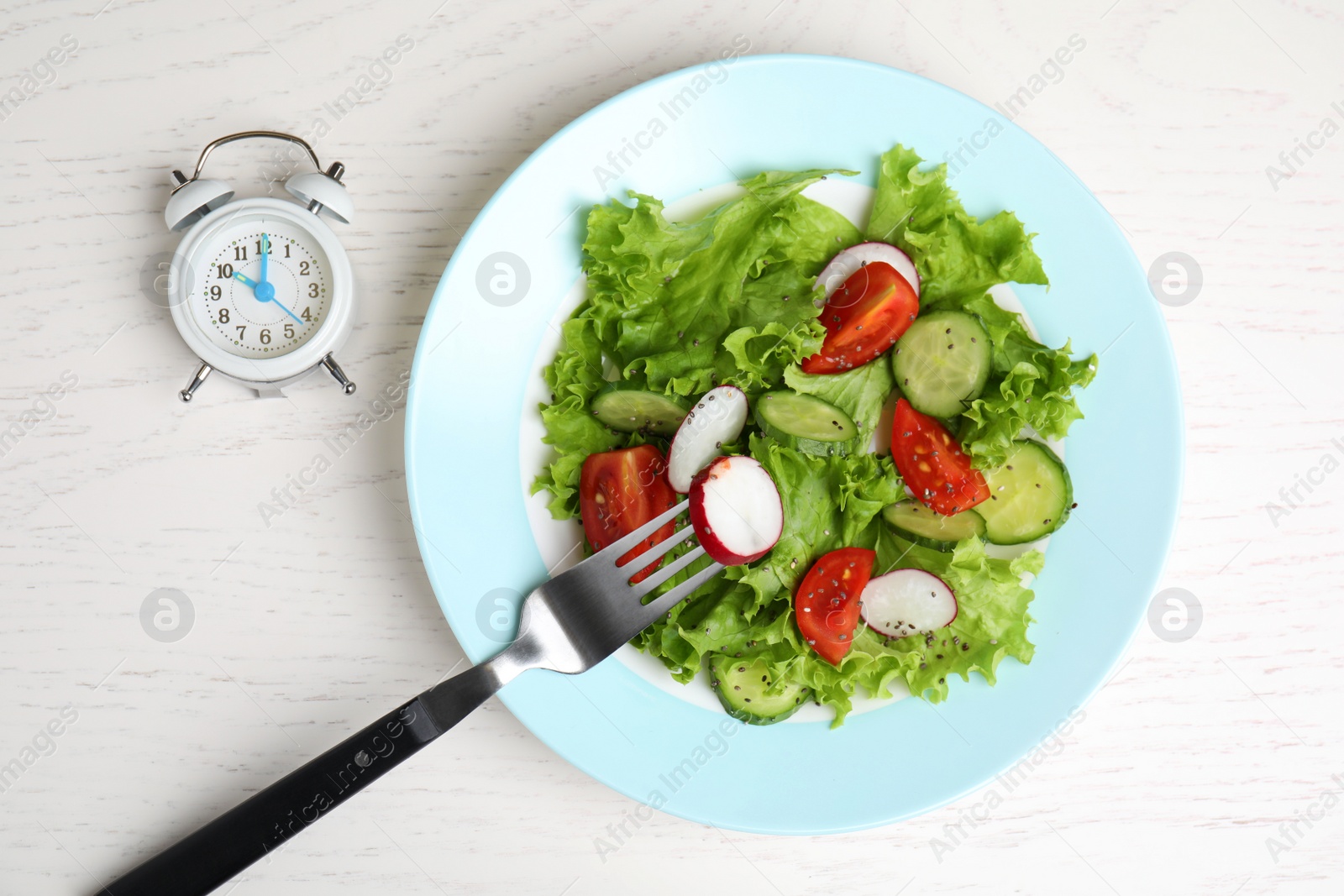 Photo of Alarm clock and plate with salad on white wooden 
table, flat lay. Meal timing concept