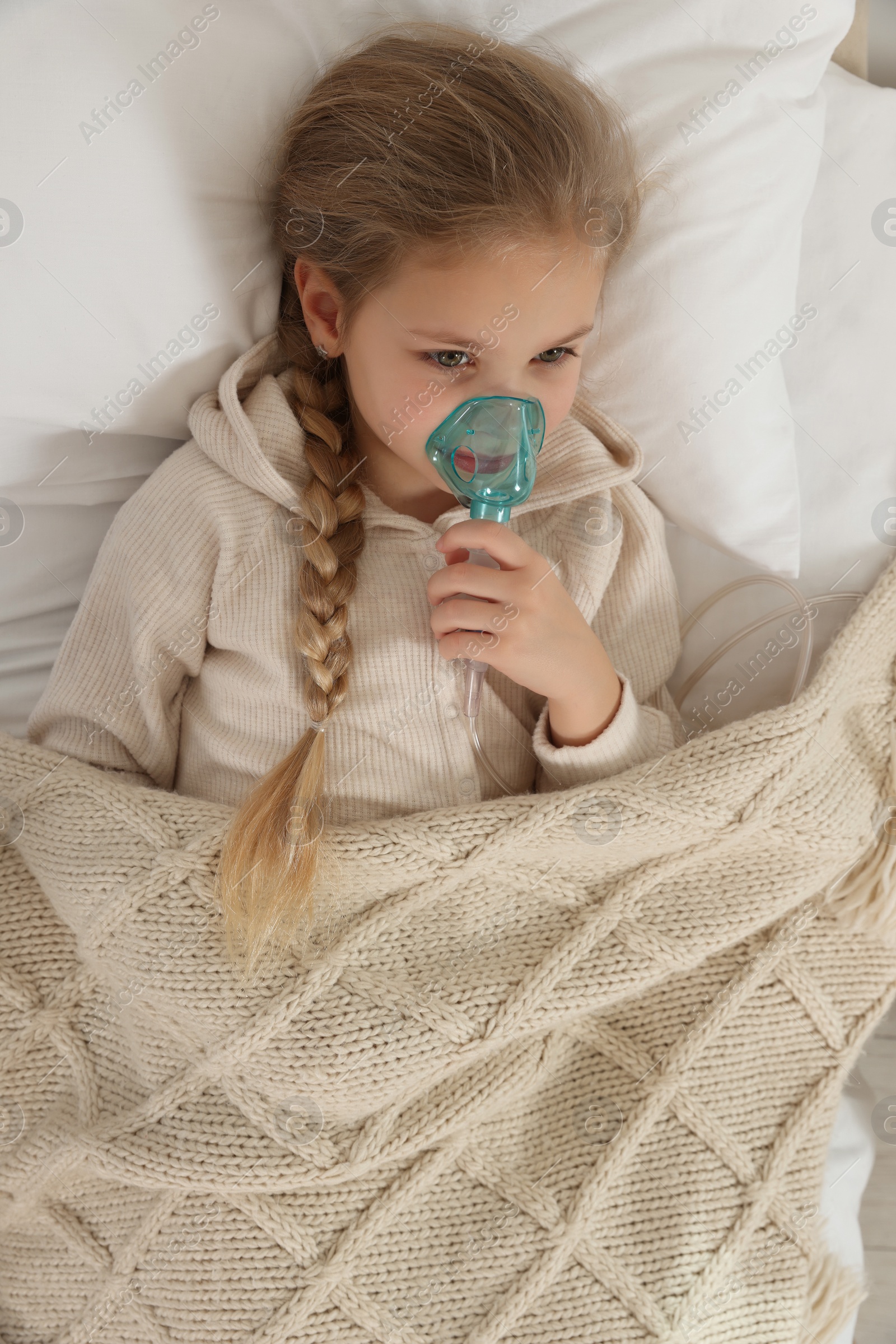 Photo of Little girl using nebulizer for inhalation on bed indoors, top view