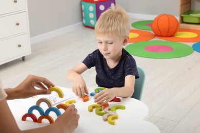 Motor skills development. Mother helping her son to play with colorful wooden arcs at white table in room