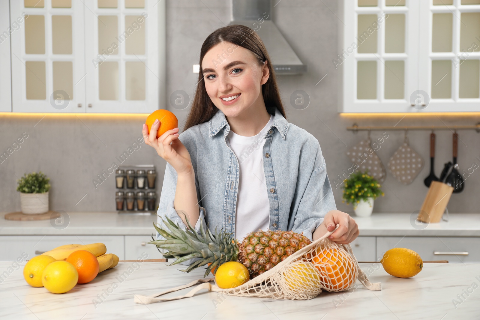 Photo of Woman with string bag of fresh fruits at light marble table in kitchen