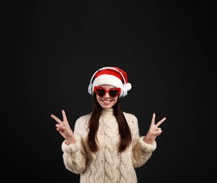 Young woman in Santa hat listening to Christmas music on black background