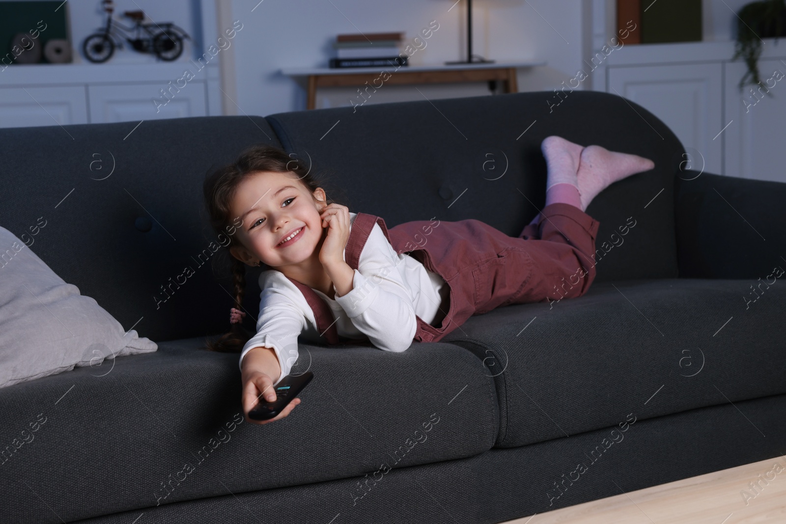 Photo of Cute little girl changing TV channels with remote control on sofa at home