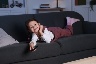 Photo of Cute little girl changing TV channels with remote control on sofa at home