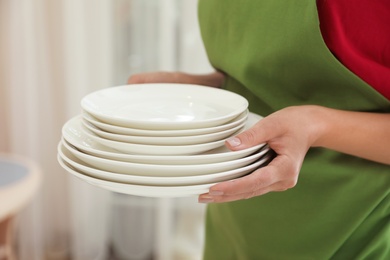 Photo of Woman holding stack of clean dishes indoors, closeup