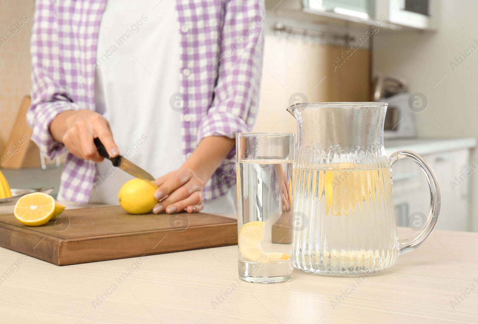 Photo of Young woman making lemon water in kitchen, focus on jug and glass with drink