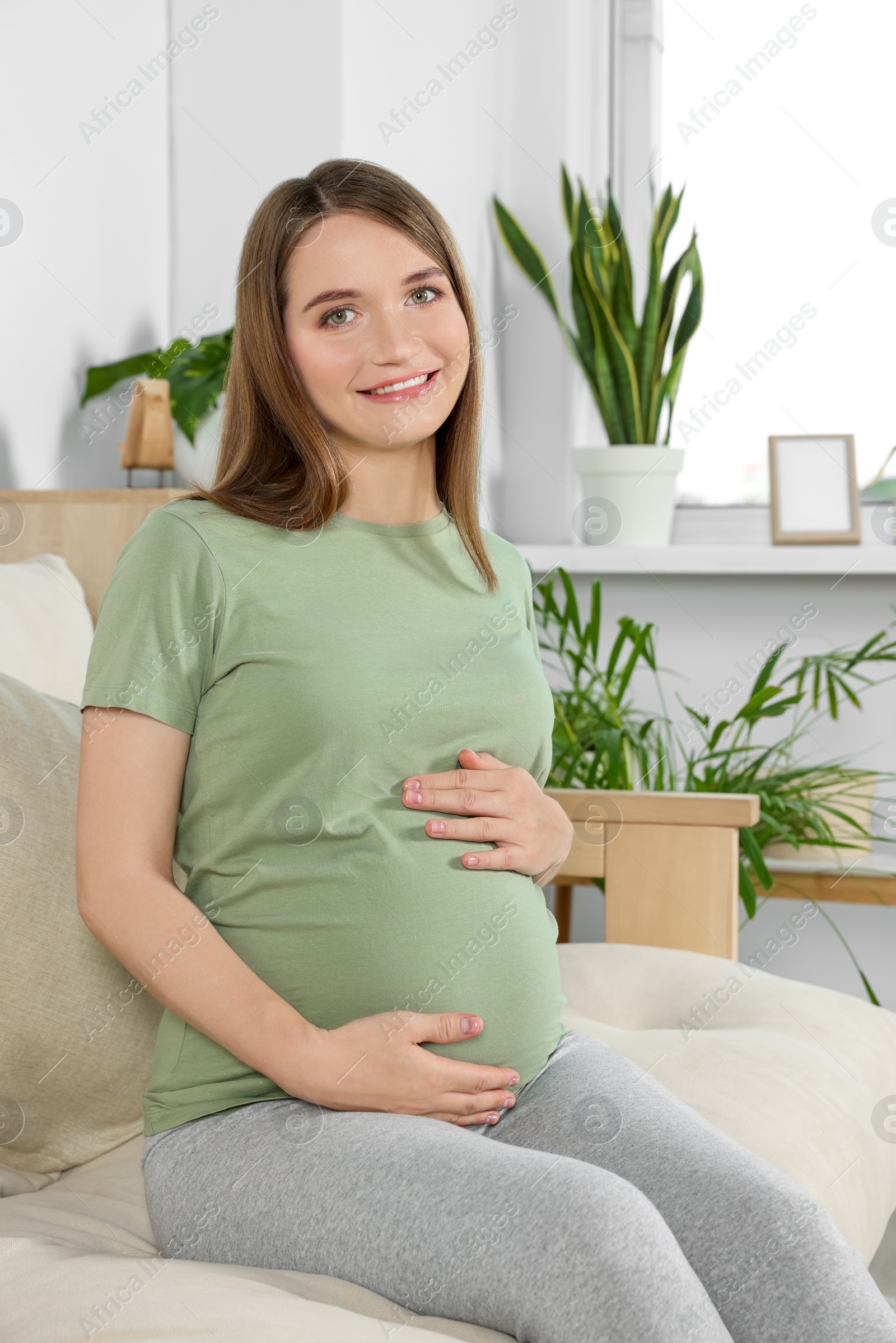 Photo of Happy pregnant woman sitting on sofa and touching her belly in living room