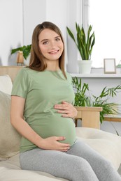 Happy pregnant woman sitting on sofa and touching her belly in living room