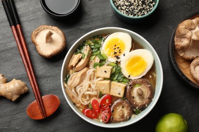 Photo of Bowl of delicious ramen, ingredients and chopsticks on black table, flat lay. Noodle soup