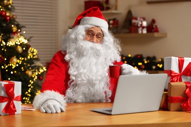 Photo of Santa Claus with cup of drink. Laptop and Christmas gifts on table at home