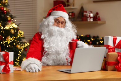 Santa Claus with cup of drink. Laptop and Christmas gifts on table at home