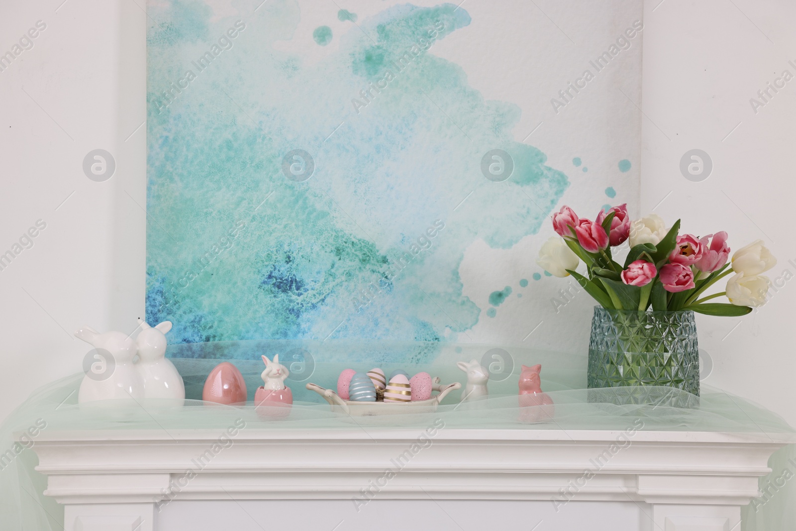 Photo of Easter decorations. Bouquet of tulips in vase, painted eggs and bunny figures on fireplace at home