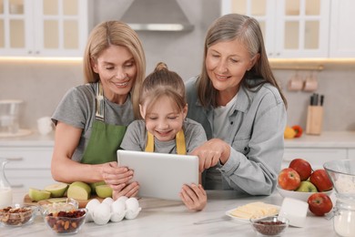 Photo of Three generations. Happy grandmother, her daughter and granddaughter using tablet in kitchen