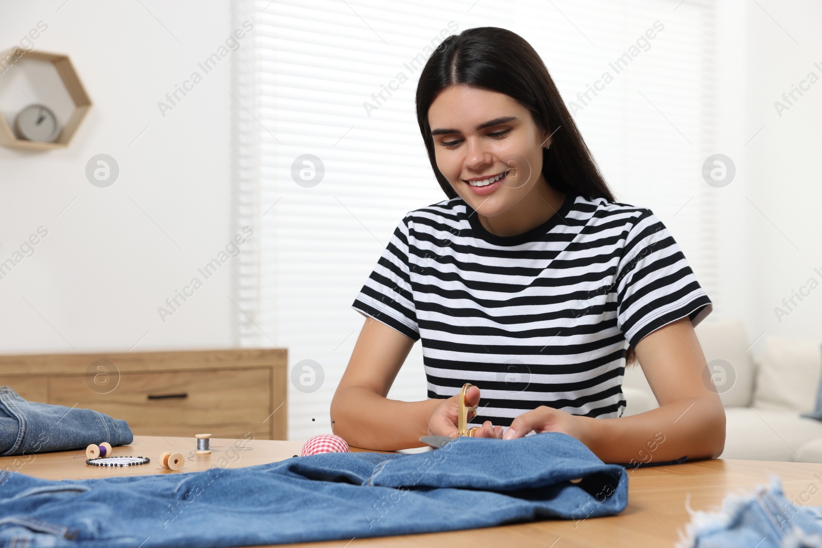 Photo of Young woman shortening jeans with scissors at wooden table indoors, space for text