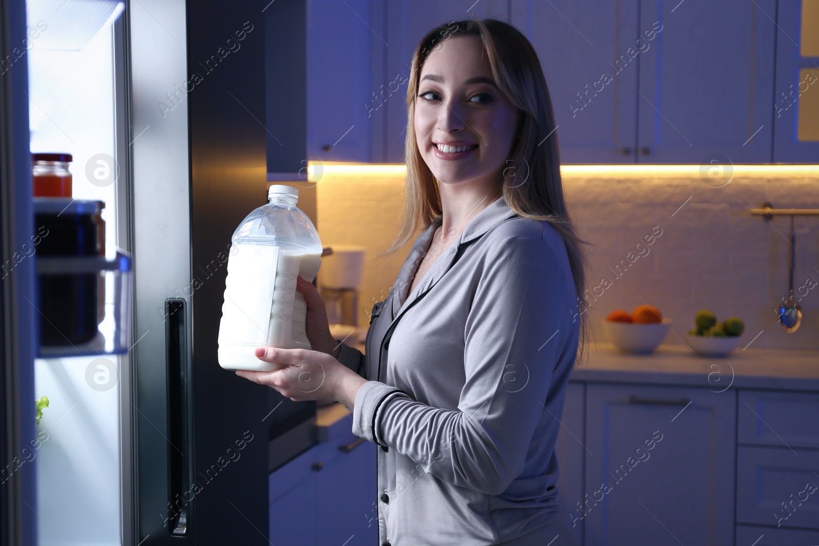 Photo of Young woman holding gallon bottle of milk near refrigerator in kitchen at night