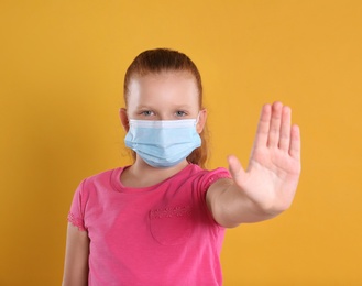 Photo of Little girl in protective mask showing stop gesture on yellow background, space for text. Prevent spreading of coronavirus