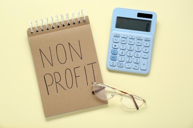 Photo of Notebook with phrase Non Profit, glasses and calculator on beige background, flat lay