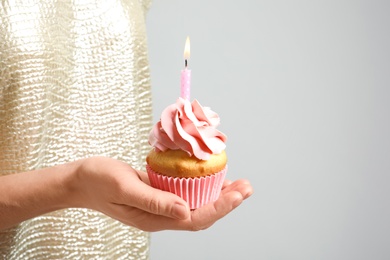 Photo of Woman holding delicious birthday cupcake with burning candle on light background, closeup