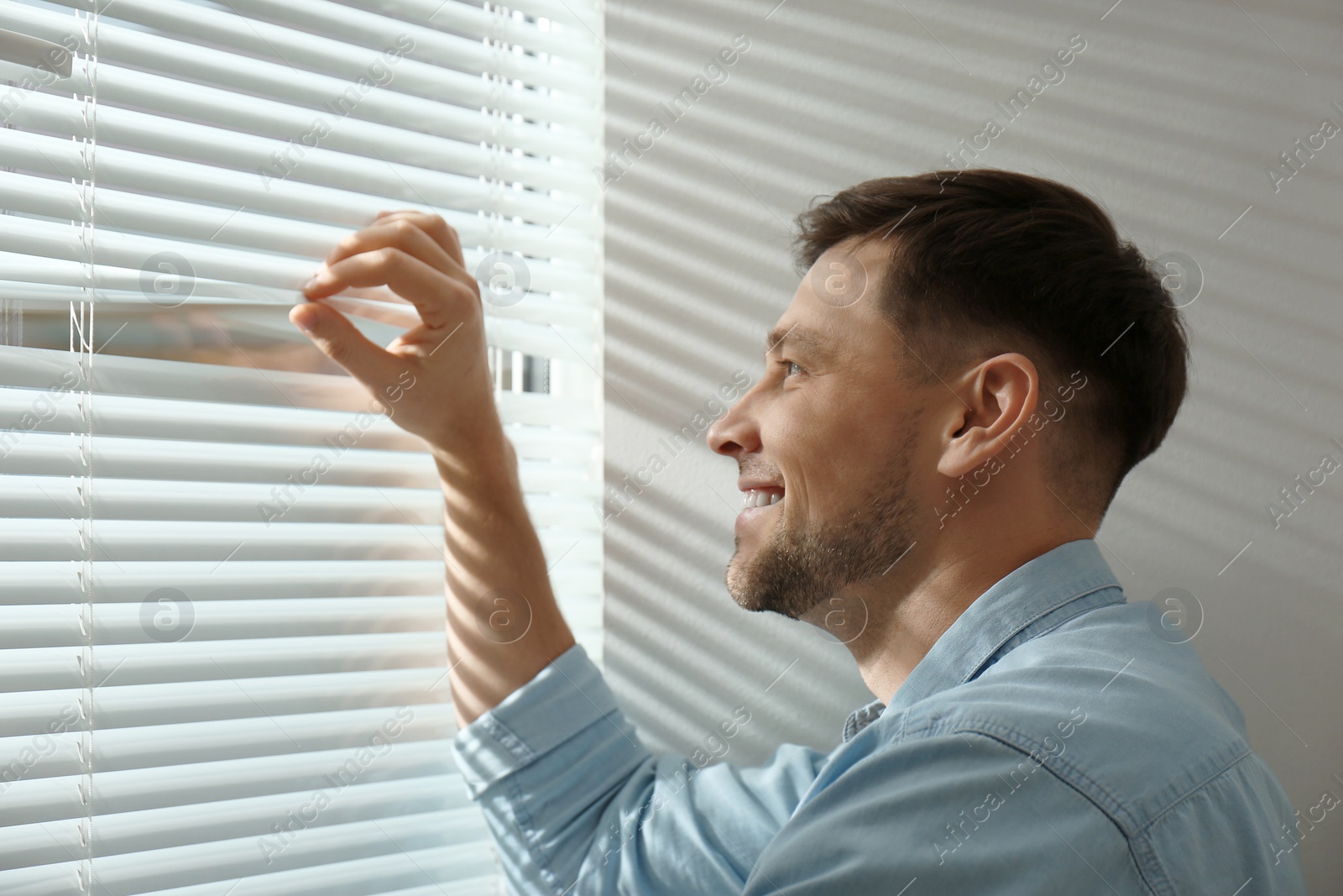 Photo of Handsome man opening window blinds at home