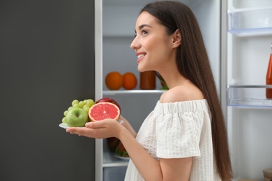 Photo of Concept of choice between healthy and junk food. Woman holding plate with fruits near refrigerator in kitchen