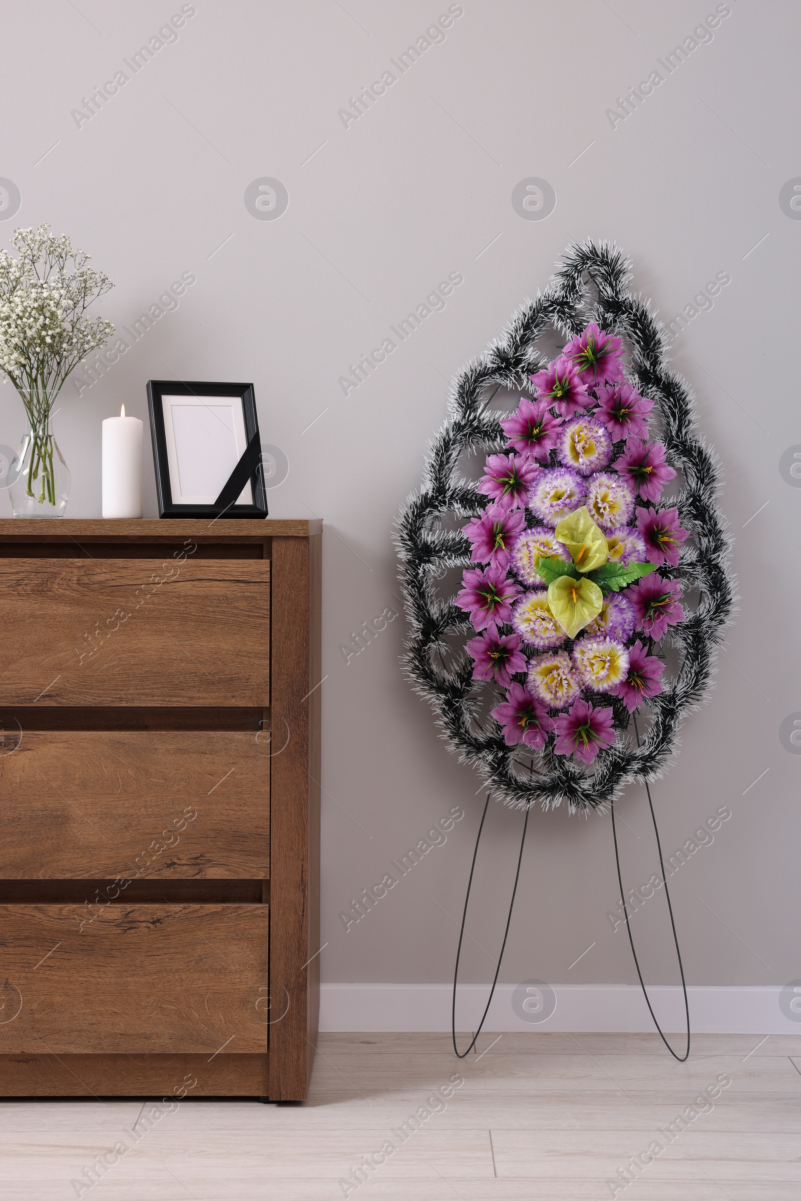 Photo of Wreath of plastic flowers and frame with black ribbon, burning candle on commode in room. Funeral attributes