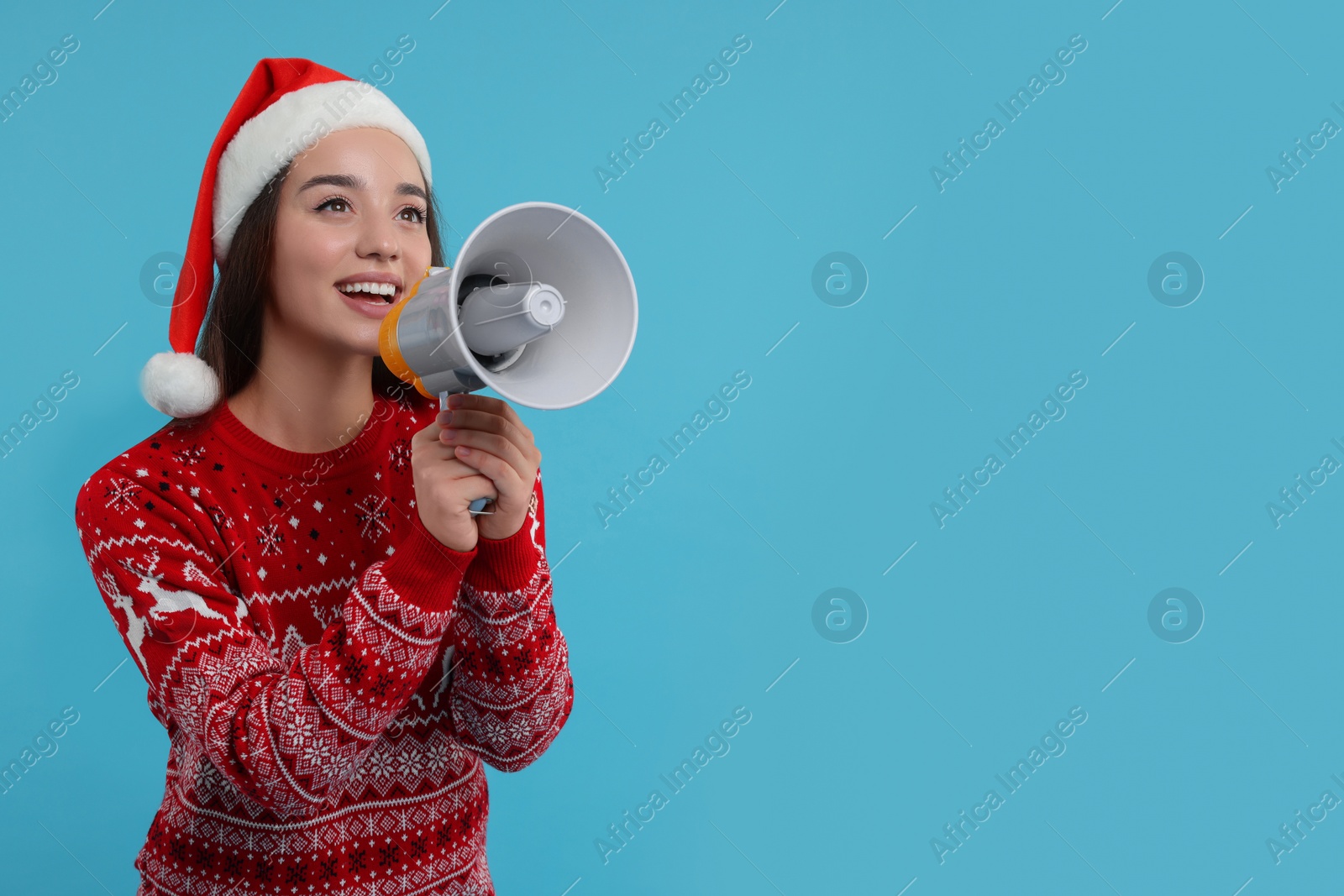 Photo of Young woman in Christmas sweater and Santa hat shouting in megaphone on light blue background. Space for text