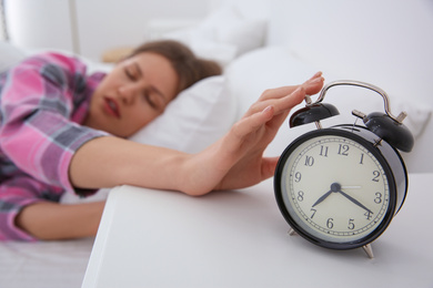 Photo of Sleepy young woman turning off alarm clock at home in morning, focus on hand