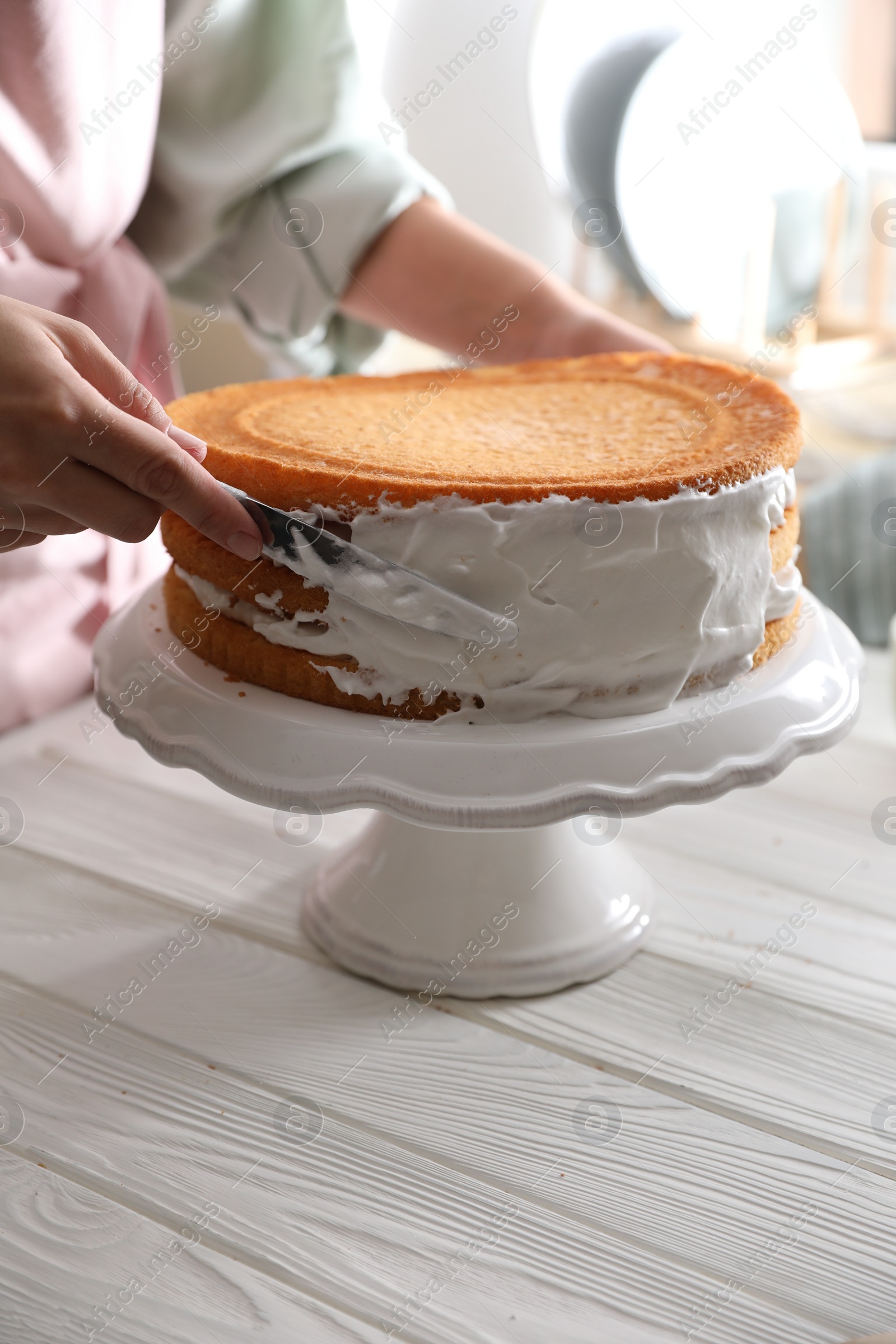 Photo of Woman smearing sides of sponge cake with cream at white wooden table, closeup