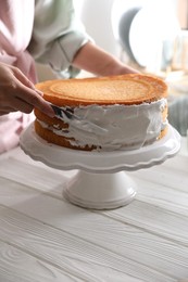 Photo of Woman smearing sides of sponge cake with cream at white wooden table, closeup