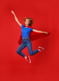 Photo of Confident woman in superhero cape jumping on red background