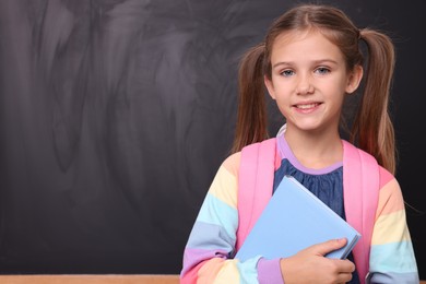 Photo of Smiling schoolgirl with book near blackboard. Space for text