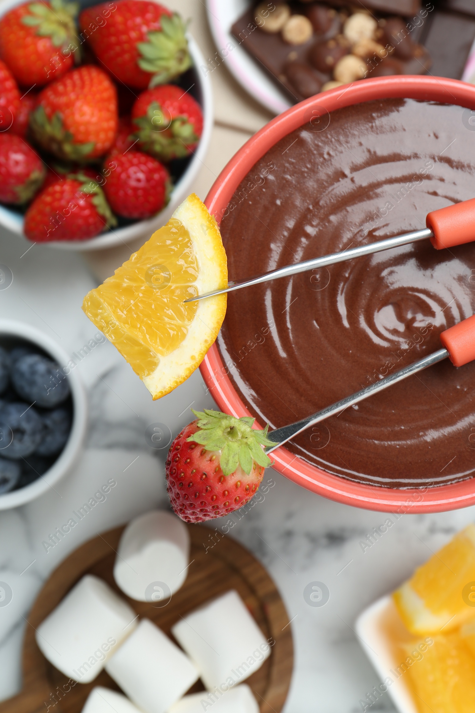 Photo of Fondue pot with melted chocolate, marshmallows, fresh orange, different berries and forks on white marble table, flat lay