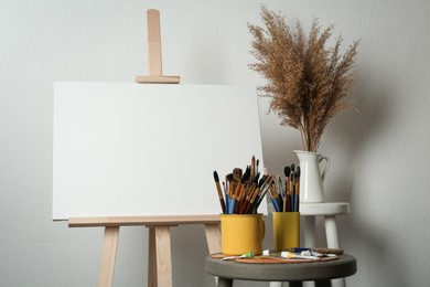 Photo of Easel with blank canvas, brushes, paints and palette in studio