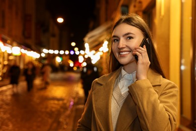 Photo of Smiling woman talking by smartphone on night city street. Space for text