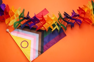 Photo of Rainbow paper garland and bright progress flag on orange background, flat lay. LGBT pride