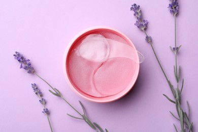 Photo of Under eye patches in jar with spatula and lavender flowers on lilac background, flat lay. Cosmetic product