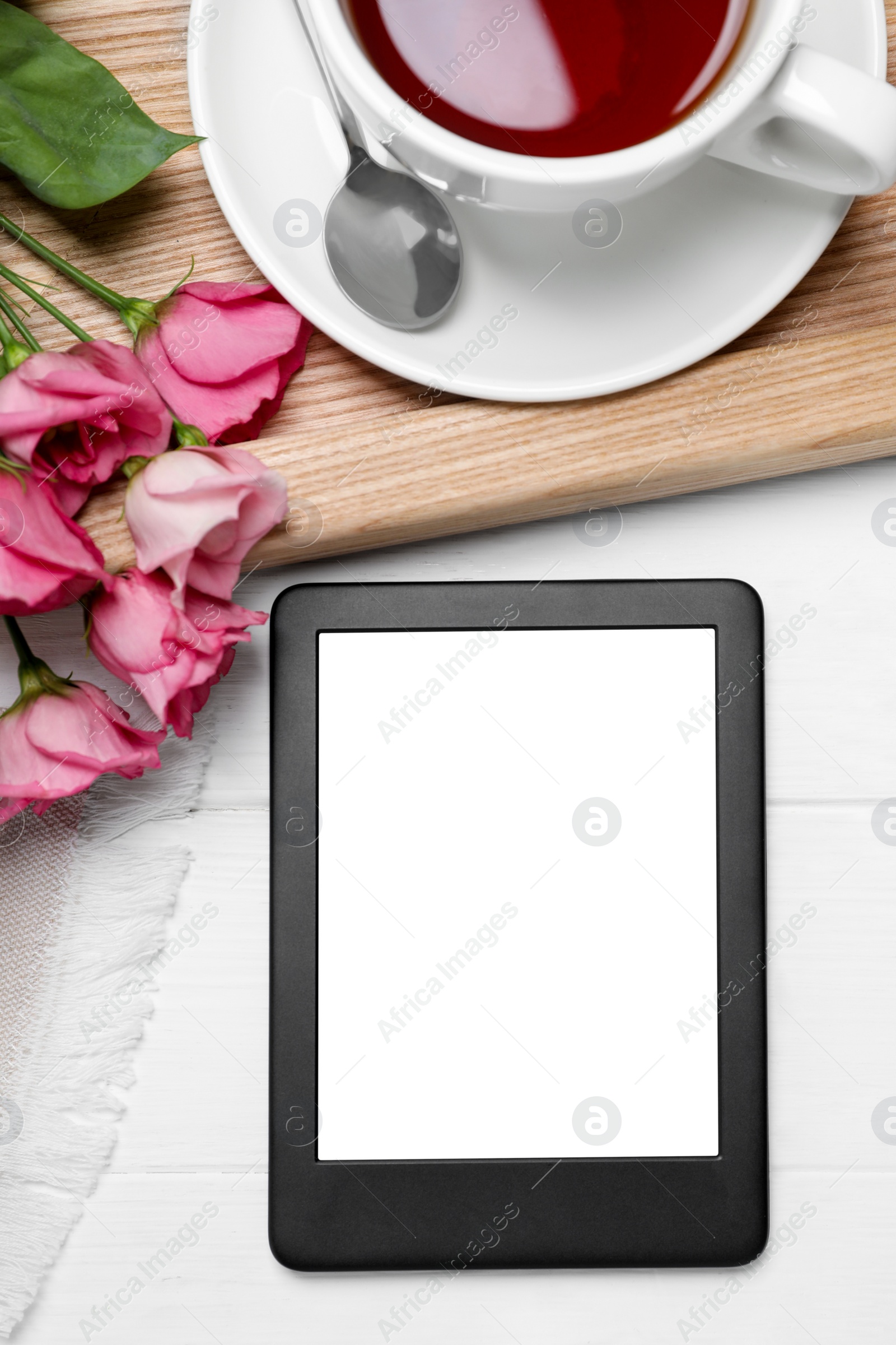 Photo of Modern e-book reader near cup of tea and flowers on white wooden table, flat lay