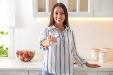 Photo of Young woman holding glass of pure water in kitchen, focus on hand
