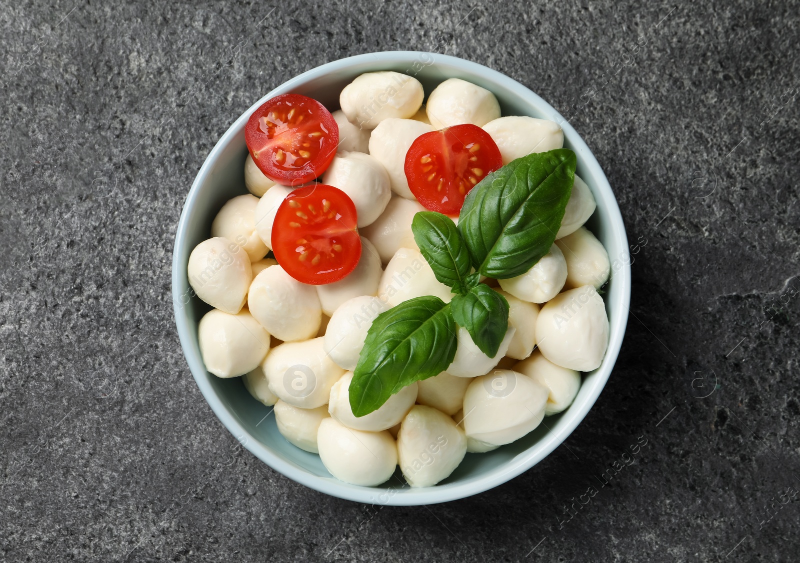 Photo of Delicious mozzarella balls, tomatoes and basil leaves in bowl on light gray textured table, top view