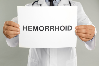 Doctor holding sign with word HEMORRHOID on light grey background, closeup