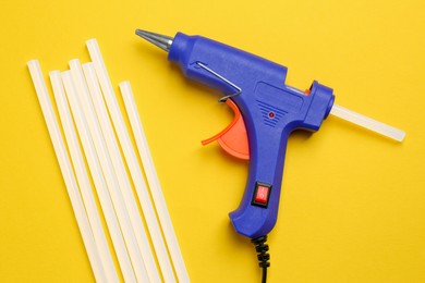 Photo of Blue glue gun and sticks on yellow background, flat lay